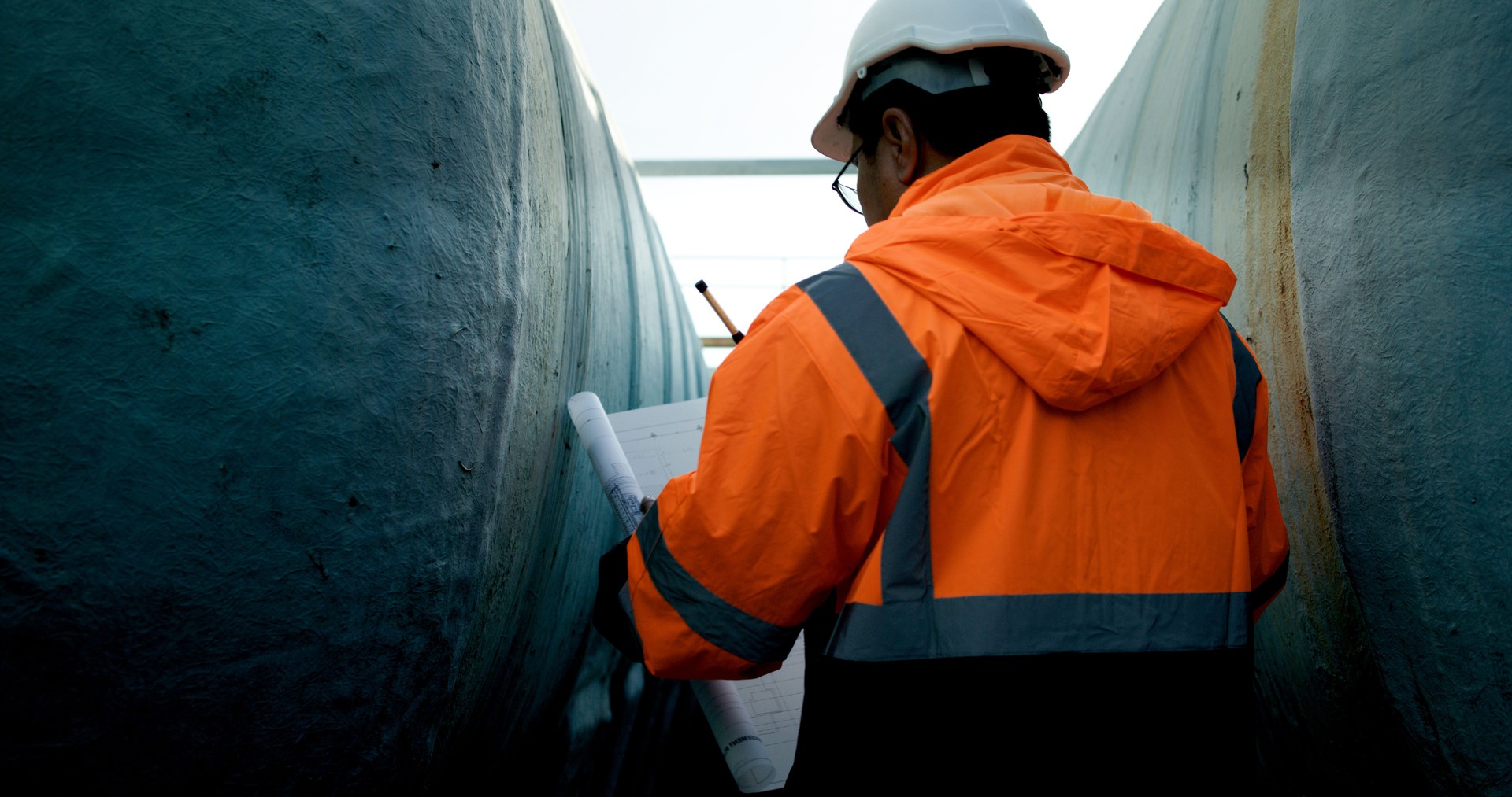 Mechanical Engineer wearing orange safety jacket and white hardhat with laptop  and blueprint working on horizontal water tanks at construction site